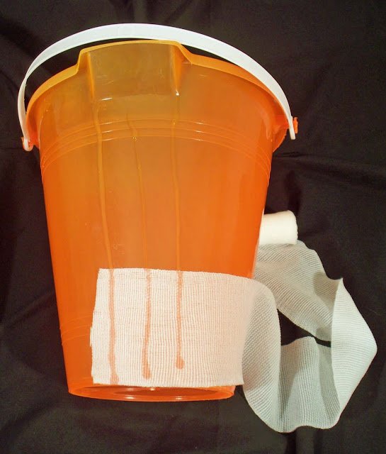 orange bucket with beads of glue and gauze being wrapped around it to make a mummy bucket. 
