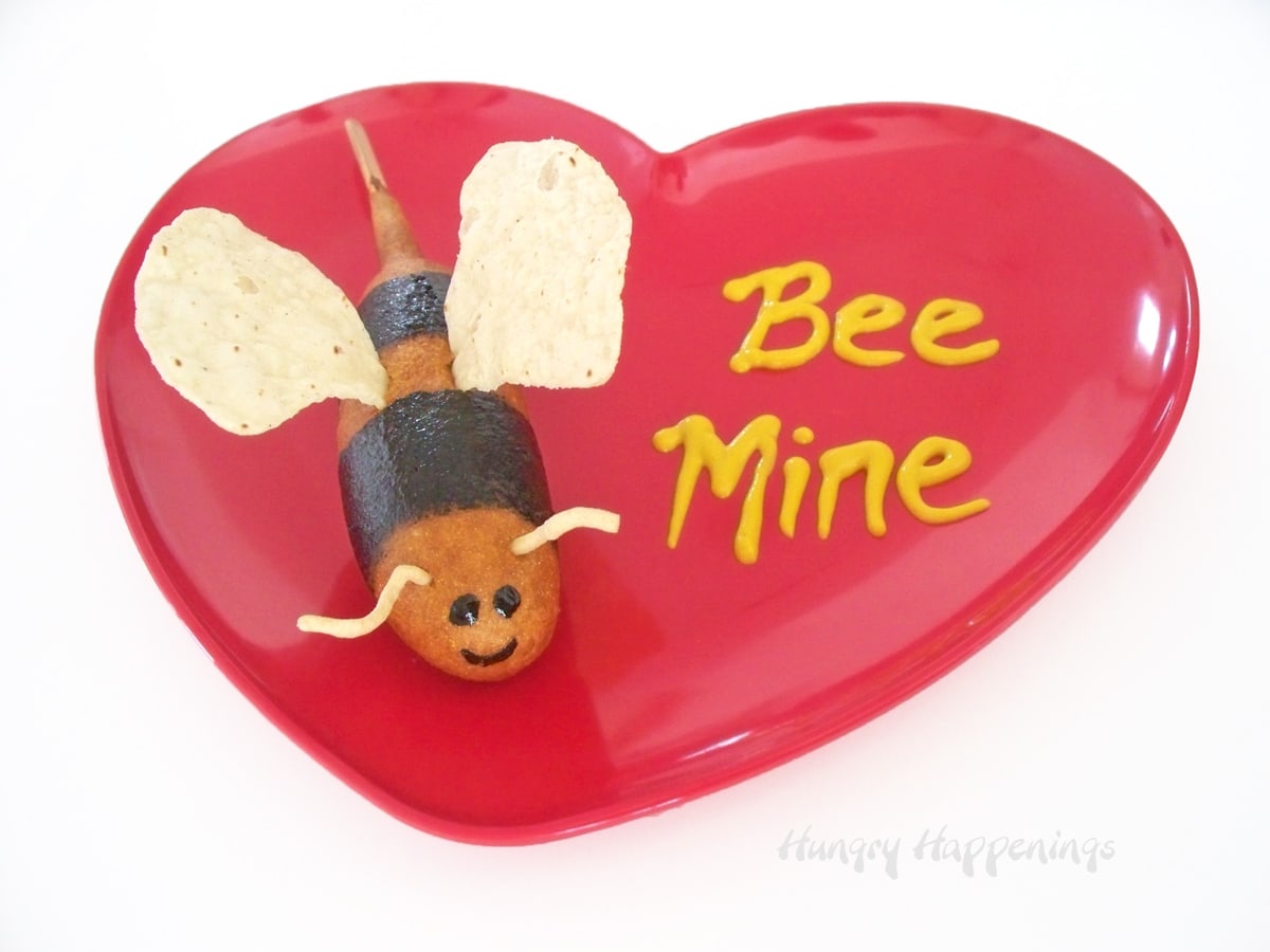 bumble bee corn dog served on a red heart plate with "bee mine" printed in mustard. 