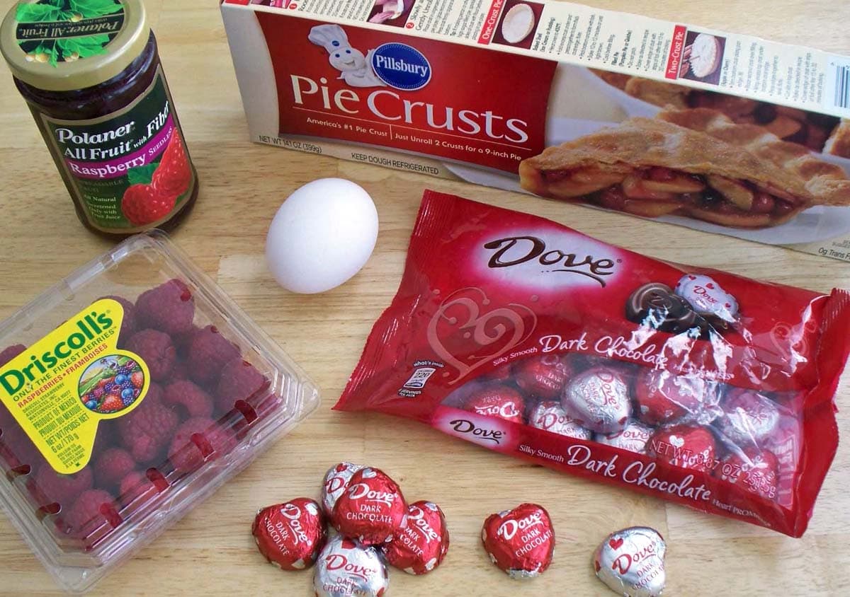 chocolate pastry hearts ingredients including pie dough, raspberry jelly, chocolate hearts, an egg, and raspberries.