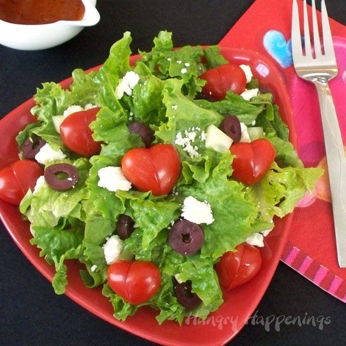 fresh salad greens topped with heart-shaped tomatoes, feta cheese, cucumbers, and Kalamata olive slices served on a red plate