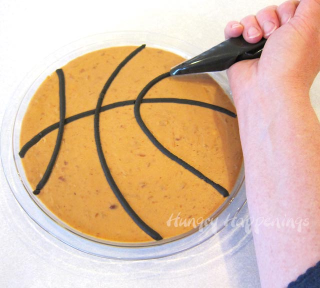 piping seam lines onto a basketball bean dip using black refried beans