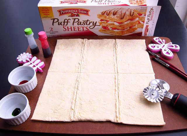 puff pastry unrolled and cut into 6 rectangles next to food coloring, foam stamps, paintbrushes, and a pizza cutter. 