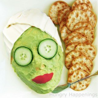 cheese ball shaped like a woman's face covered in a green guacamole mask, cucumber eyes, red pepper lips, and mozzarella head wrap
