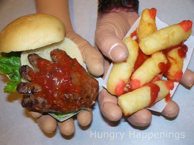 Hand-shaped hamburger served on a bun with blood-red ketchup and a side of finger-shaped French fries with red fingernails and more ketchup blood. 