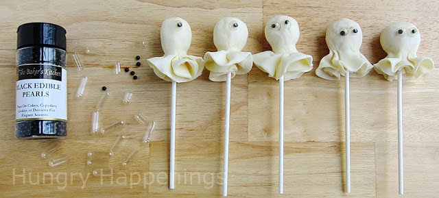 adding edible googly eyes made with gel capsules and black sugar pearls to ghost lollipops.