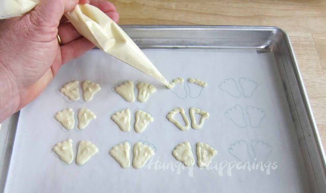 piping white chocolate over the warm fuzzy feet template that is set under a piece of parchment paper on a baking sheet.