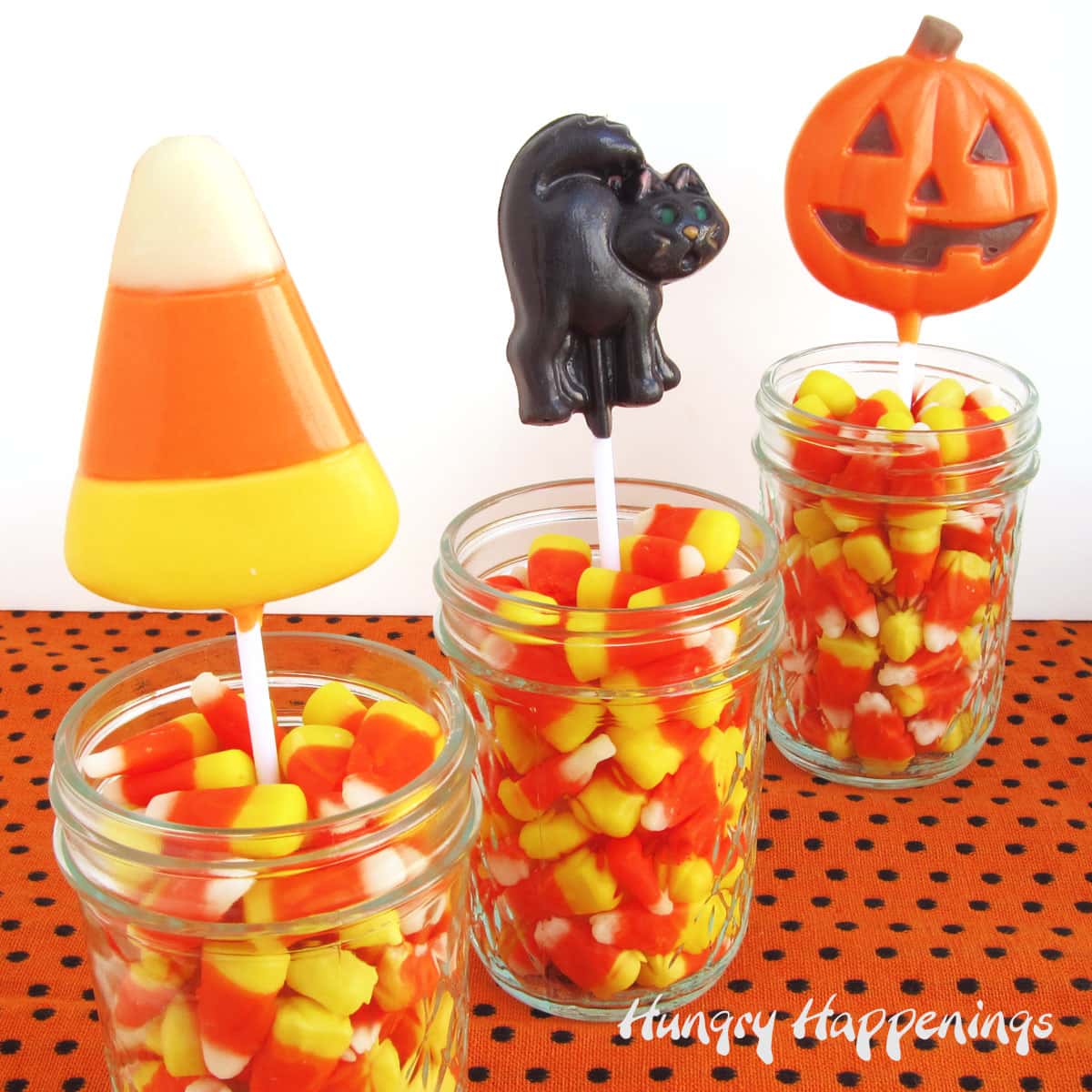 Halloween candy corn, black cat, and pumpkin lollipops displayed in jars of candy corn.