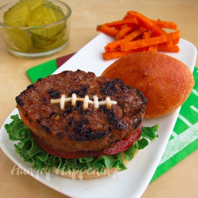 football bun topped with lettuce, tomato, and a football burger on a plate with sweet potato fries next to a bowl of pickles. 