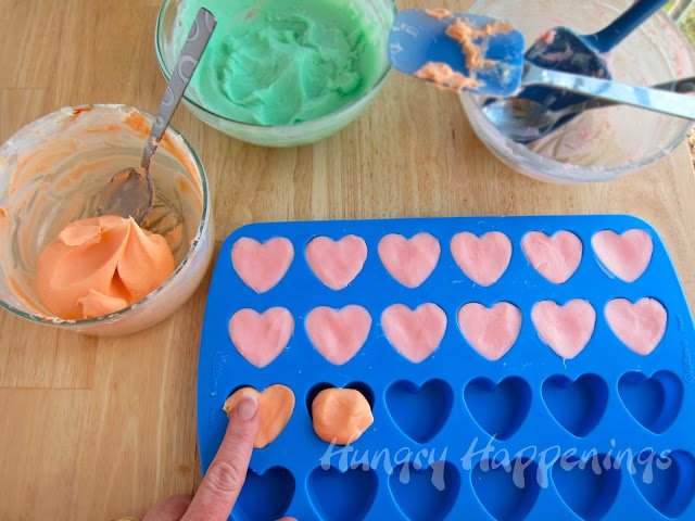 Filling heart-shaped silicone mold with pink and orange fudge. The mold is next to a bowl of orange fudge and a bowl of green fudge. 