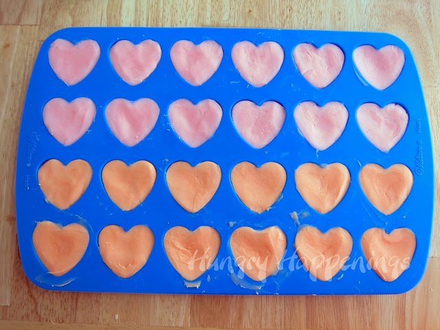 A blue silicone heart mold with 24 cavities is filled with orange and pink-colored white chocolate fudge. 