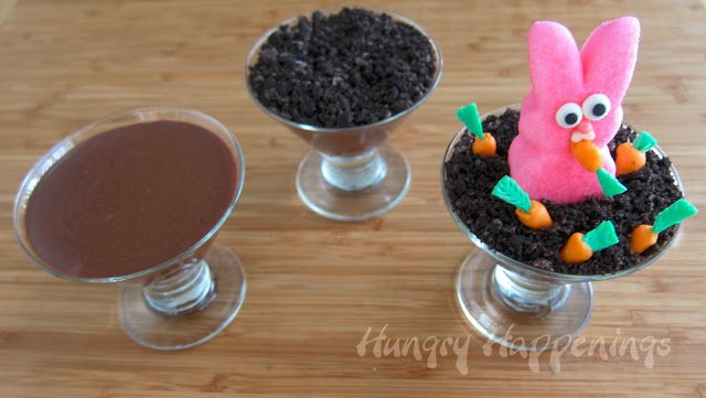three glass dessert cups, one filled with chocolate pudding, one with cookie crumbs on top, and one with a Peeps bunny eating candy carrots. 