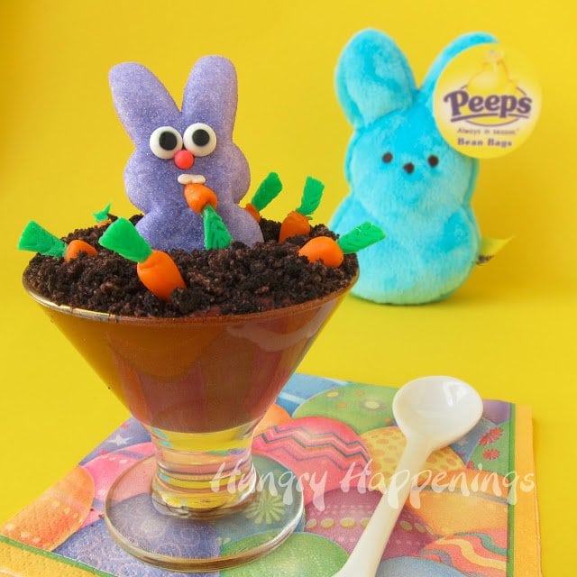 a clear glass dish filled with chocolate pudding and topped with cookie crumb dirt and candy carrots with a purple Peeps Bunny in the middle is set on an Easter napkin near a stuffed Peep bunny. 