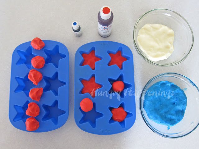 two silicone star molds being filled with red fudge next to bowls of white and blue fudge and bottles of red and blue food coloring. 