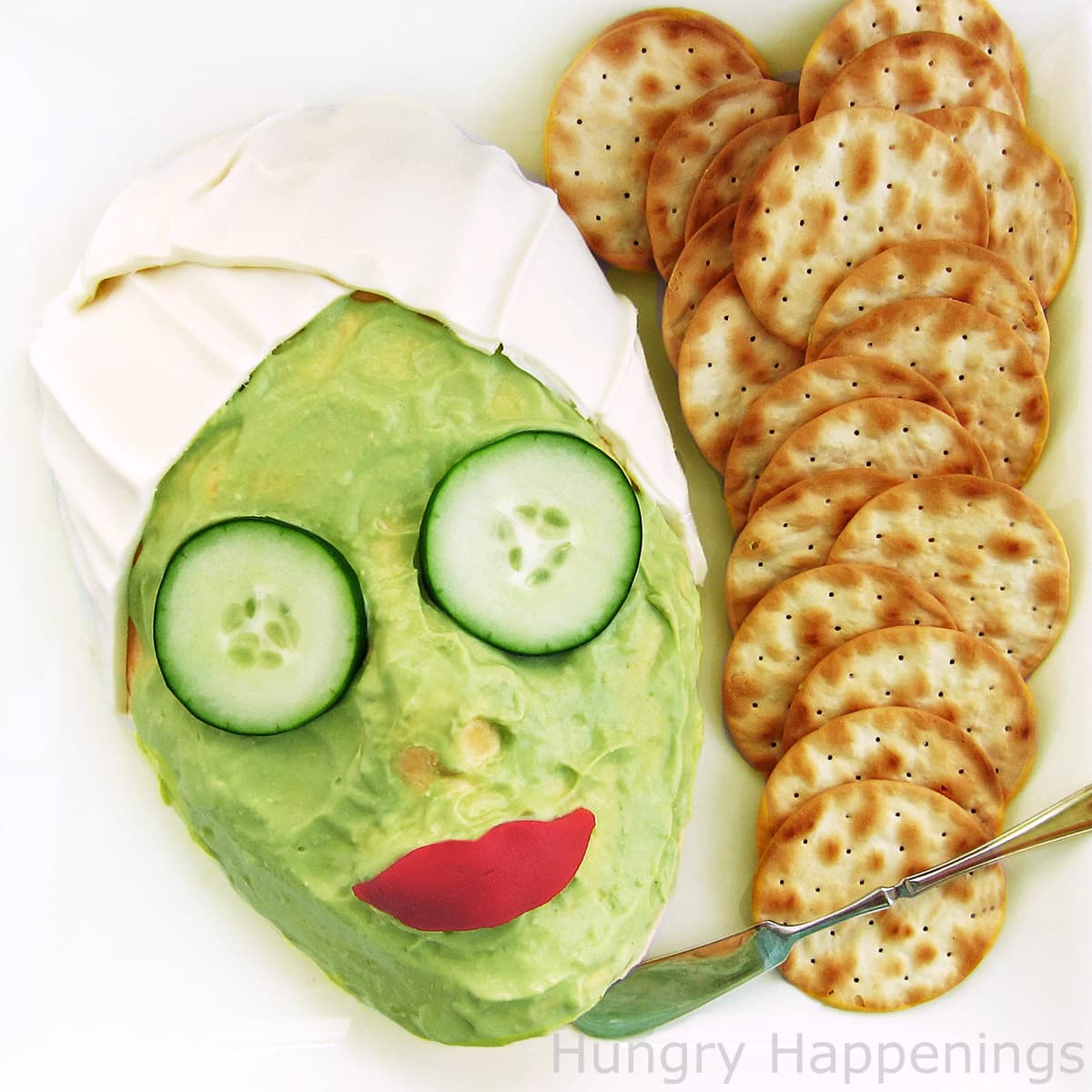 Spa party appetizer - cheese ball face covered in a mozzarella cheese wrap, a guacamole face mask, cucumbers on the eyes, and red pepper lips. Served with crackers.