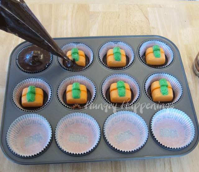 piping chocolate cake batter into a cupcake wrapper that is filled with 2 mini cheesecake pumpkins. 