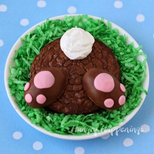 a small round brownie decorated with a frosting tale and mini Reese's Cup paws is set on green coconut grass on a white dish on a blue polka dot paper. 