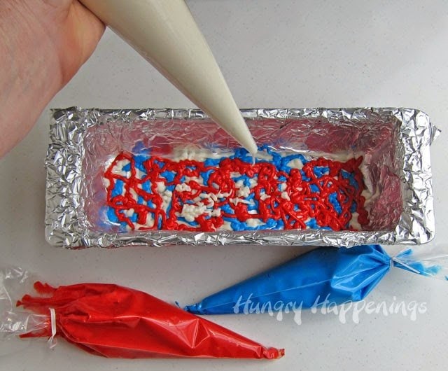 Piping white cake batter into a tin-foil lined baking pan filled with swirls of red and blue cake batter. 