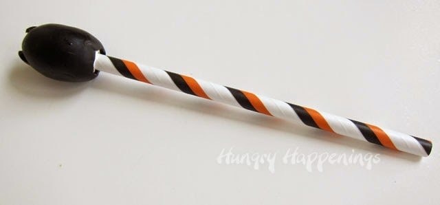 a cream cheese-filled jumbo black olive with a orange, black, and white striped paper straw inserted.