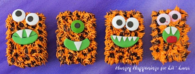 four orange and black Halloween Rice Krispie treat monsters in a row on a purple watercolor background. 
