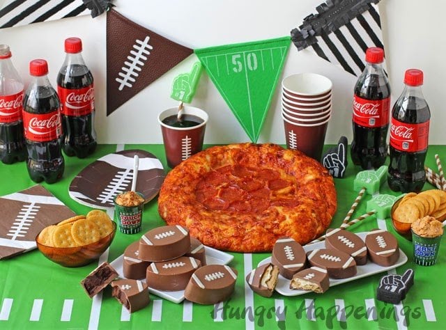 Super Bowl party food table with pennants, football plates and napkins, Coca Cola, pizza, and chocolate footballs. 