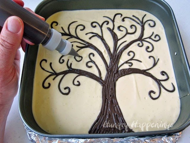piping chocolate branches onto the chocolate tree on a square cheesecake