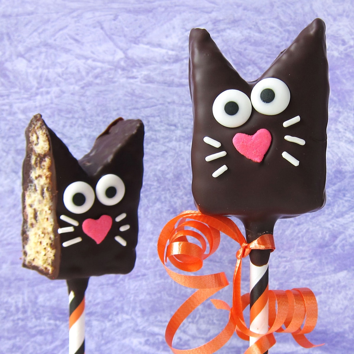 Rice Krispie Treat cats coated in dark chocolate and decorated with candy eyes, pink heart-shaped noses, and sprinkle whiskers.