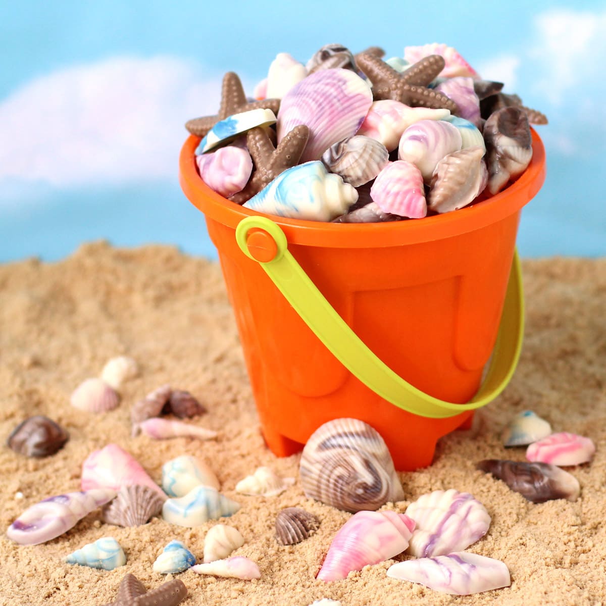 chocolate shells in an orange beach pail set on cookie crumb sand surrounded by more seashell chocolate.