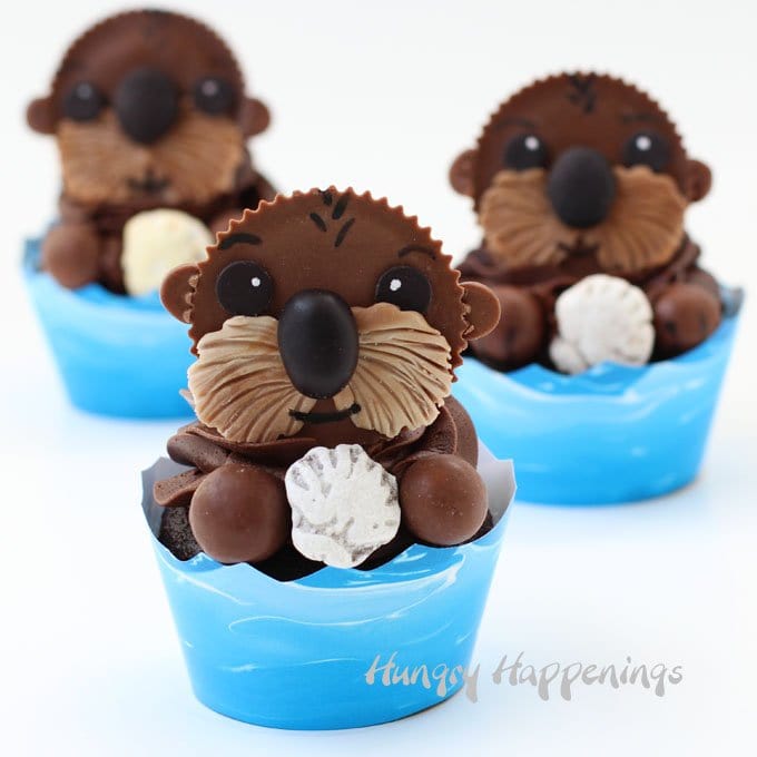 3 Reese's Cup Sea Otters holding candy shells are each on a chocolate cupcake that is wrapped in a blue ocean paper wrapper.