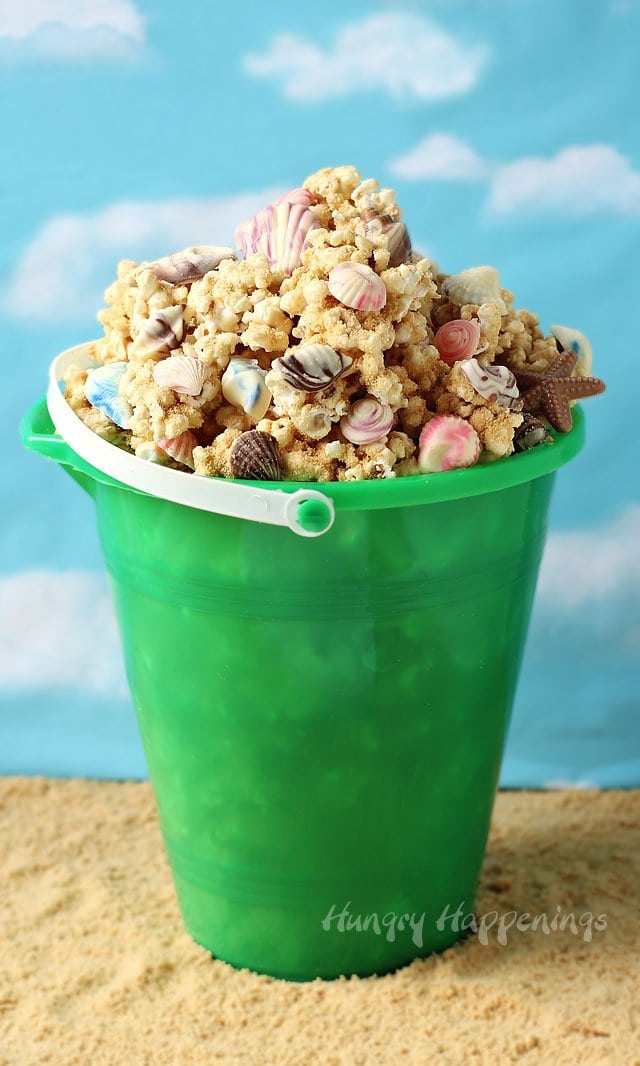 green beach pail filled with peanut butter popcorn speckled with chocolate sea shells displayed on a sandy beach. 
