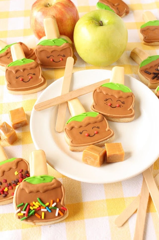 caramel apple sugar cookies decorated with royal icing set on a round white plate along with caramel squares and popsicle sticks. 