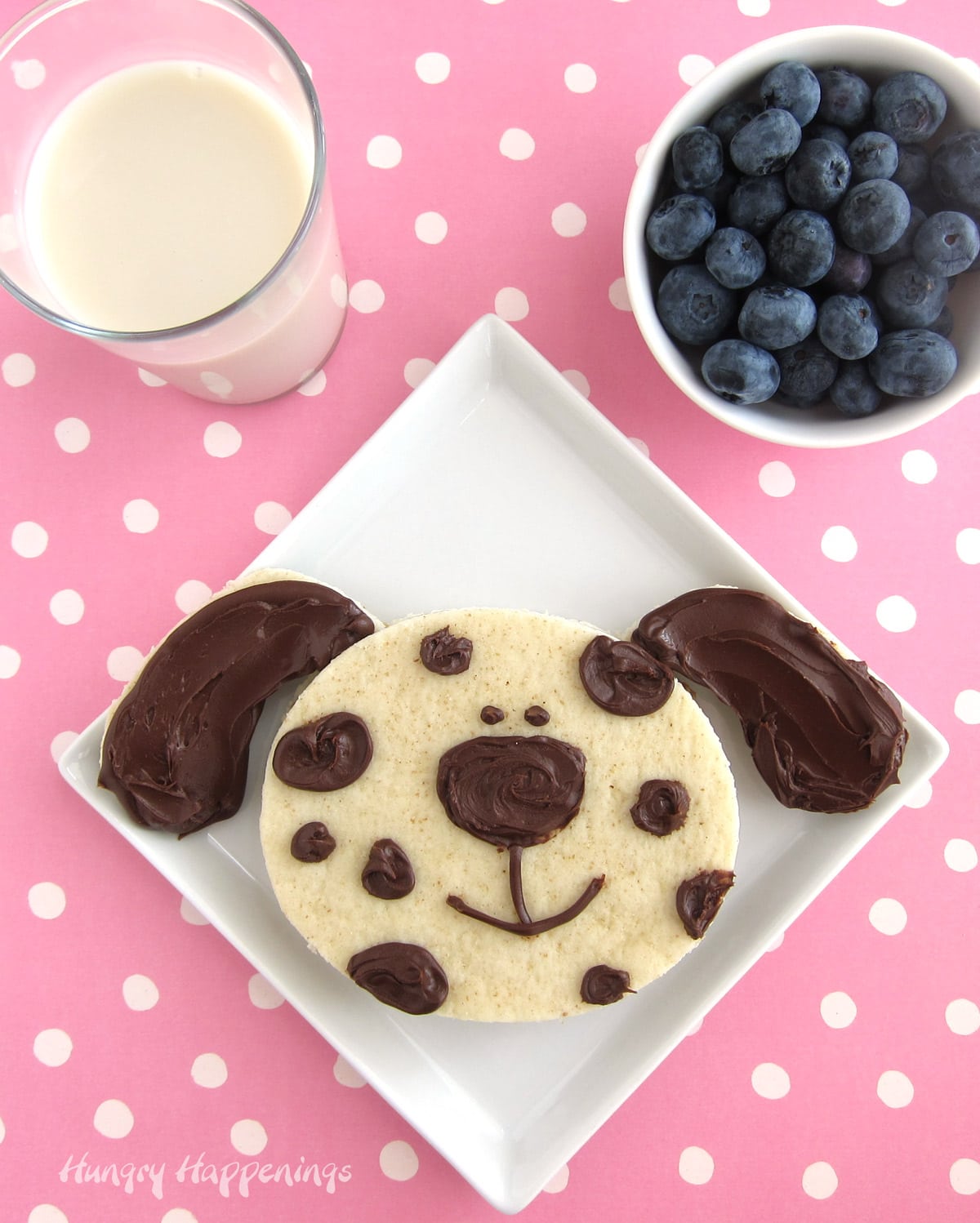 cute dog sandwich made with Nutella is served on a white plate with a glass of milk and bowl of blueberries. 