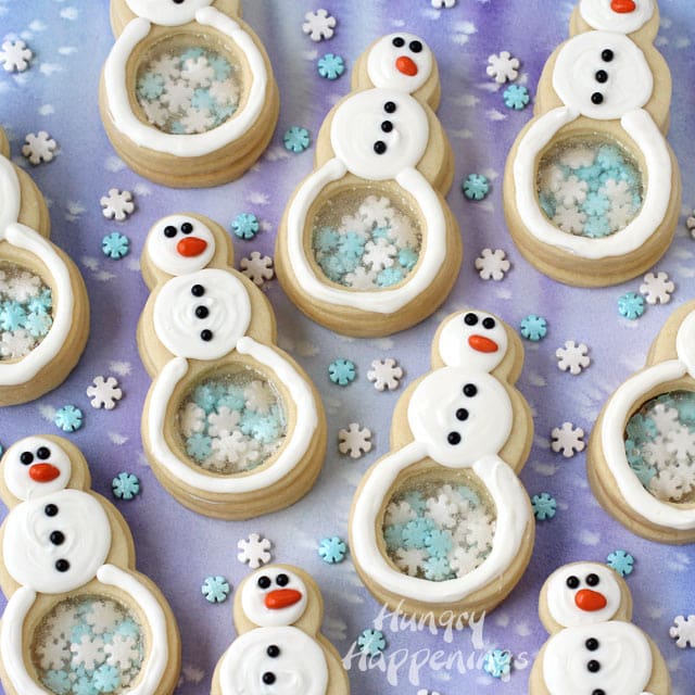 snowflake filled snowman cookies arranged on a blue watercolor background with snowflake sprinkles scattered in between the cookies