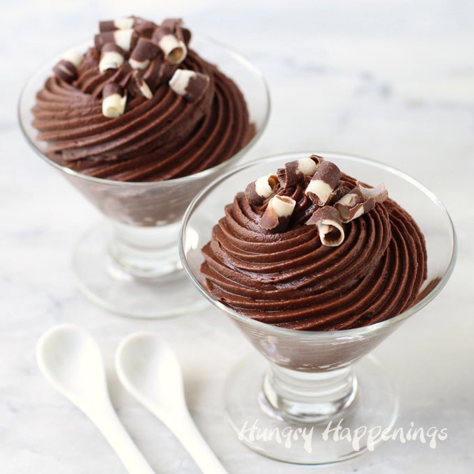 two clear cups filled with chocolate mousse each topped with dark and white chocolate curls are sitting next to two small white spoons on a marble counter top