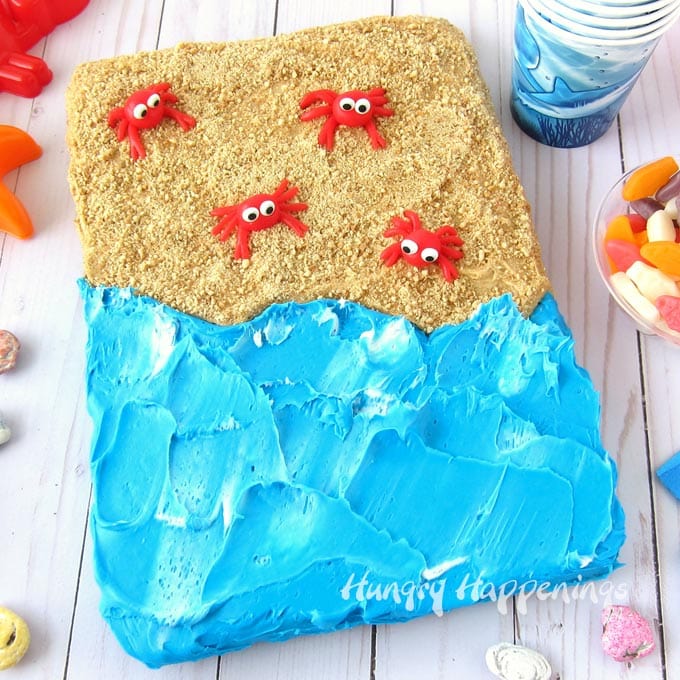 Beach cake brownie topped with peanut butter frosting and cookie crumb sand, a swirl of blue and white frosting water, and a few red candy crabs. 