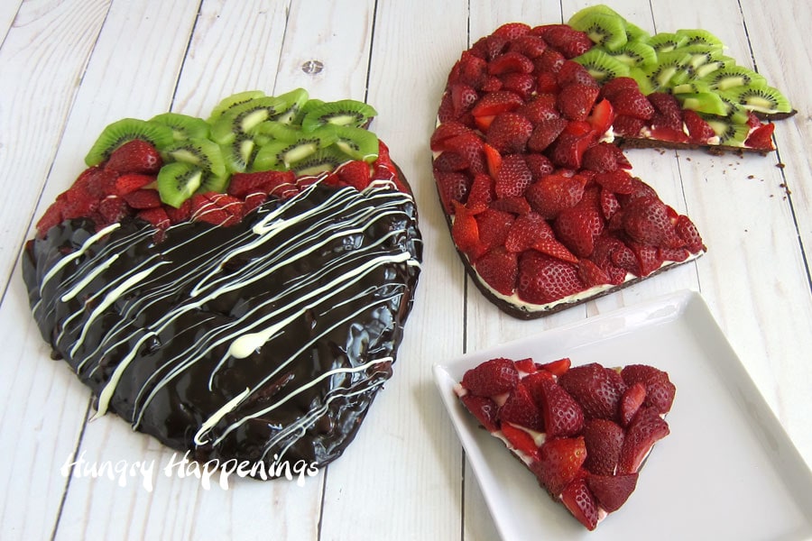 Strawberry shaped fruit pizza with a slice cut out set next to a chocolate covered strawberry fruit pizza.