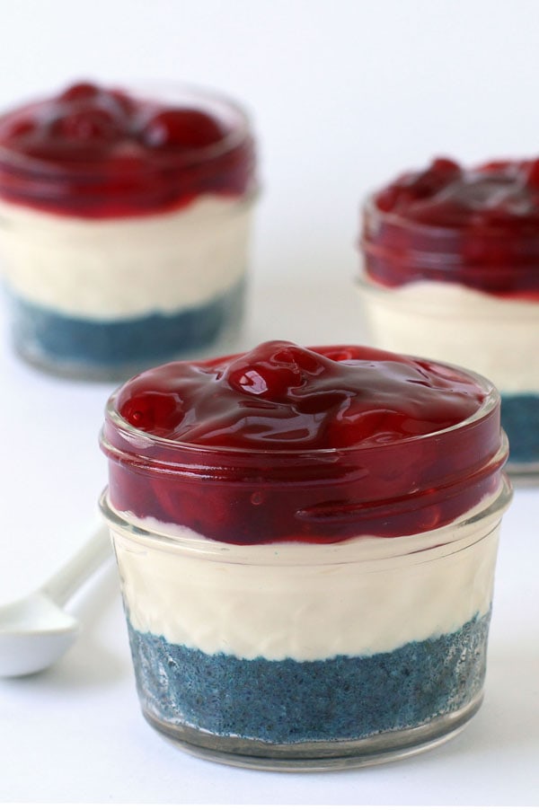 No-bake cheesecakes with a blue colored graham cracker crust and topped with cherry pie filling served in mini mason jars.
