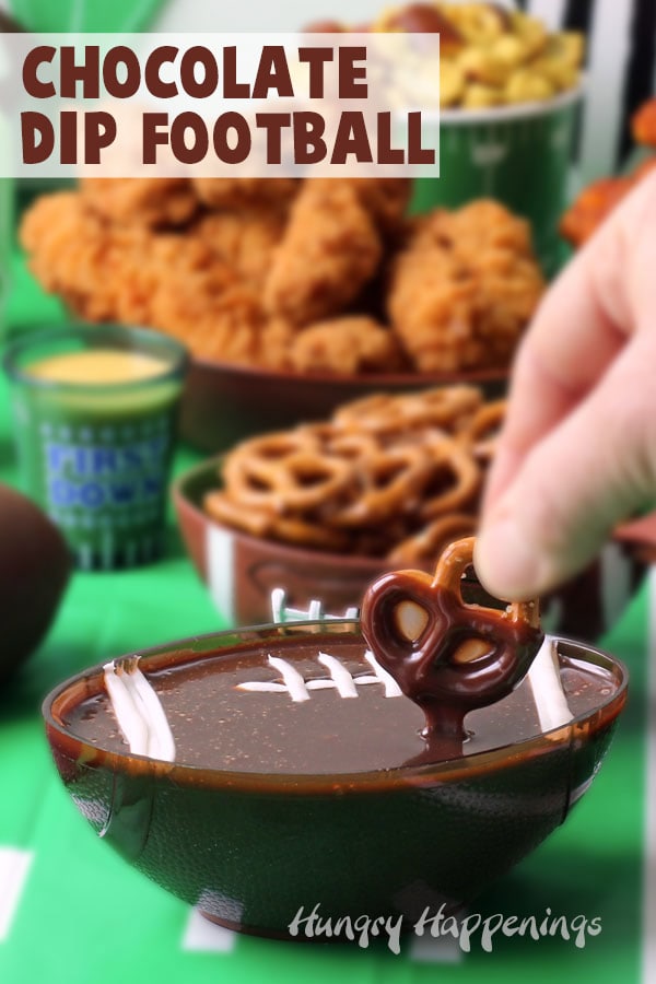 pretzel dipped into a football shaped bowl filled with chocolate ganache setting on a table along with other game day snacks