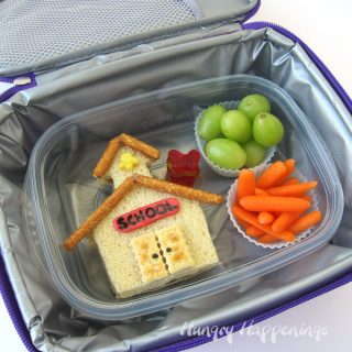 Kid's school lunch of Peanut Butter and Jelly Sandwich cut and decorated to look like a schoolhouse along with grapes and carrots.