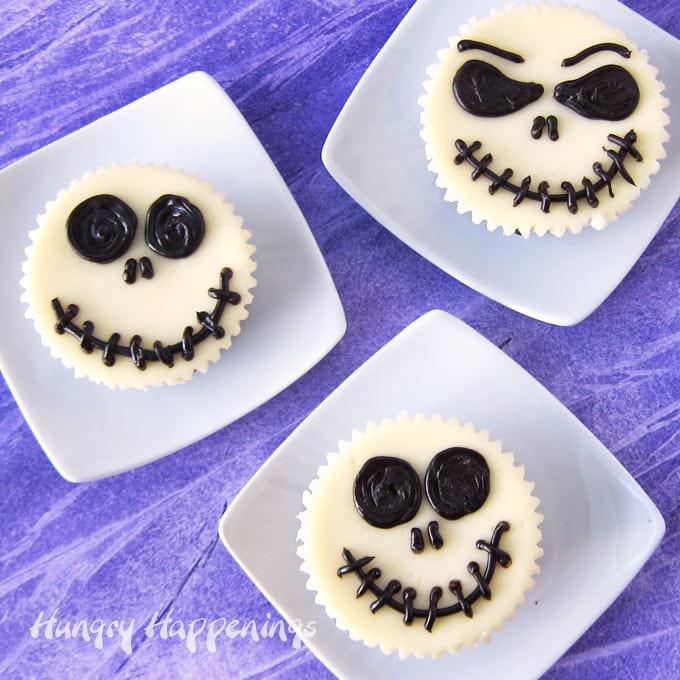 mini cheesecakes decorated to look like Jack Skellington are plated on small square white plates set on a purple watercolor backdrop