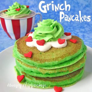 a stack of green copycake IHOP Grinch pancakes topped with cream cheese glaze, green whipped cream, and candy hearts on a white plate are set in front of a red and white striped cup filled with grinch hot chocolate