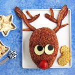 Rudolph cheese ball with bacon antlers on a white platter with bowls of star shaped and Christmas tree shaped crackers and a snowflake cheese knife all on a blue snowflake background.