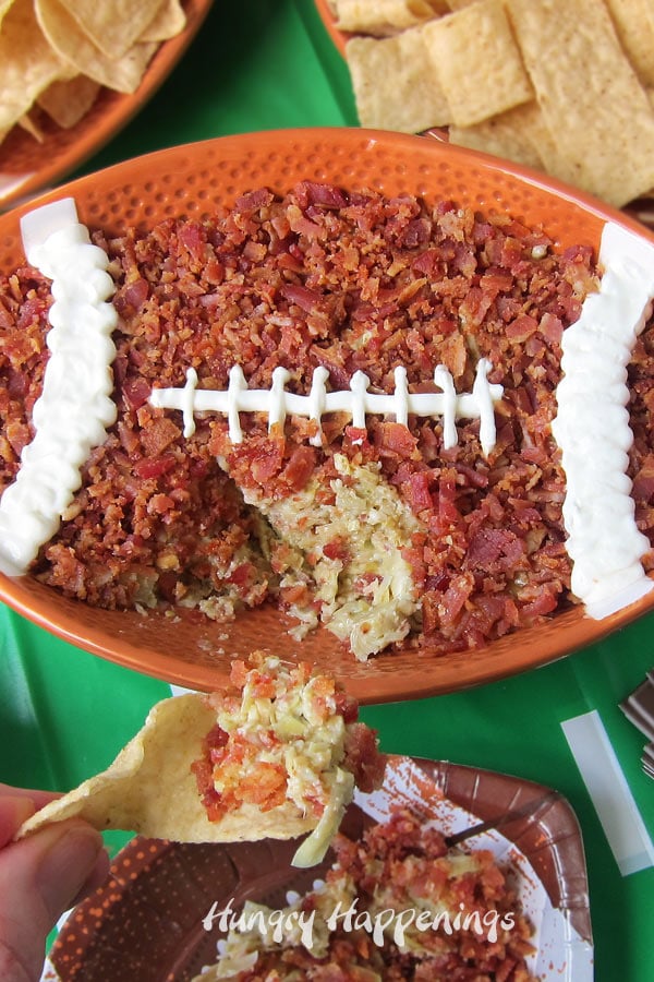 Mission Tortilla Strips topped with a scoop of the artichoke pesto dip topped with bacon that is served in a football shaped bowl. 