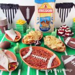Artichoke Pesto Dip served in a football shaped ceramic bowl is set in the center of a table decorated with a football field table cloth and surrounded by more football shaped plates fill with Mission Tortilla Strips, soft tacos, football topped fudge, and popcorn.