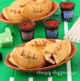 Football shapes plates topped with Calzone Footballs are set on top of a football field tablecloth along with shot glasses filled with homemade marinara sauce and tiny foam fingers and football field napkins.