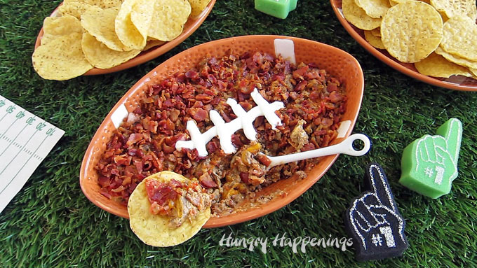 a football shaped ceramic bowl filled with hot cheeseburger dip topped with bacon and sour cream is set on Astro Turf next to football plates topped with tortilla chips and some tiny foam fingers