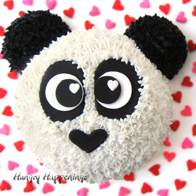 easy panda bear cake made using a round cake and two cupcakes is set on a white cake board sprinkled with candy hearts