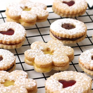 A variety of heart, daisy, and round shaped linzer cookies filled with raspberry preserves, lemon curd and Nutella are arranged on a cooling rack.