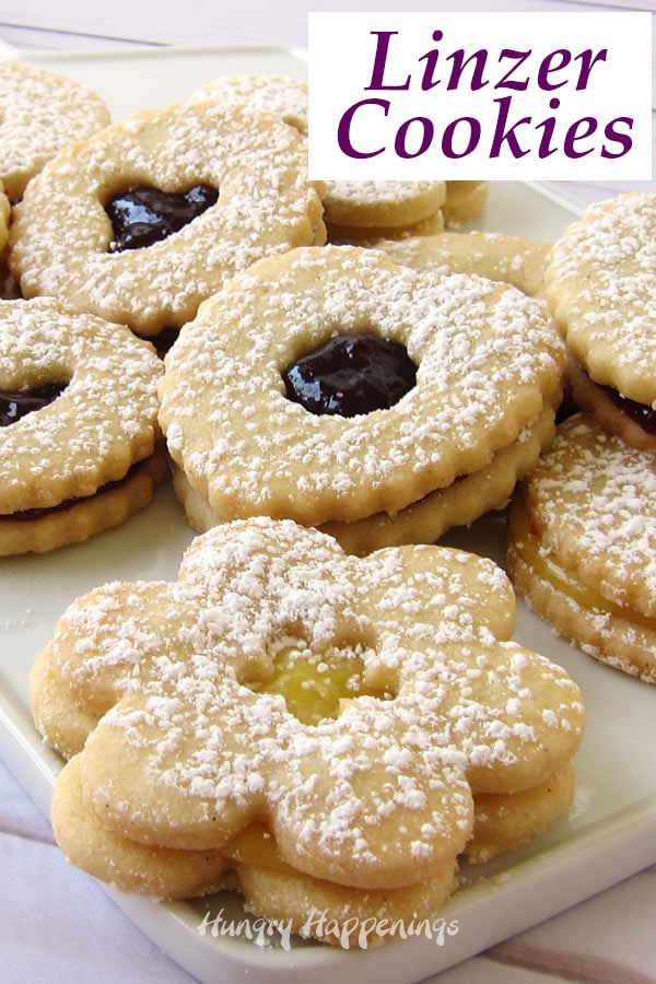 A closeup image of a variety of Linzer Cookies shaped like daisies, hearts, and rounds set on a white plate. 