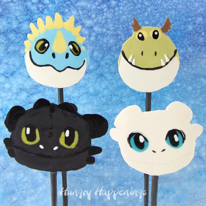 All four How To Train Your Dragon Lollipops including Night Fury (Toothless), Light Fury, Stormfly, and Meatlug, are set in front of a blue watercolor backdrop. 