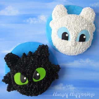 Toothless Cake and Light Fury Cake on a blue modeling chocolate covered round cake board are displayed on a blue sky watercolor background.